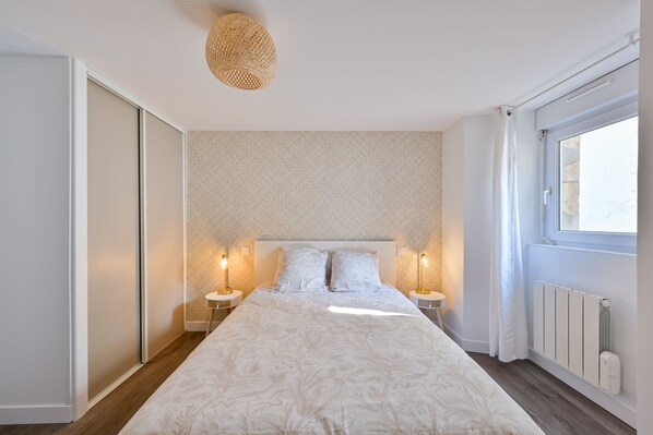 Une chambre à coucher avec un grand lit, deux tables de chevet avec des lampes, un luminaire au plafond, une fenêtre avec des rideaux et un radiateur.