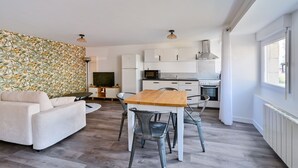 A modern kitchen with white cabinets, a wooden dining table, and a sofa in a living area with a patterned wall. - L’Escale morbihannaise – Appt à 15 min de la plage (Muzillac)
