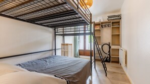 Ein Etagenbett mit Metallgestell, ein Einzelbett mit grauer Matratze, eine Holzleiter, ein Stauraum mit Regalen und eine Pendelleuchte.