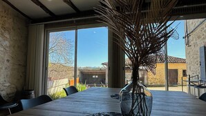 A wooden table with a vase of dried branches, large windows, and a view of a building in the background. - Villa with Pyrenees view near Foix (Cassagne)