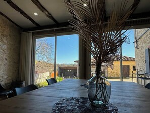 A wooden table with a vase of dried branches, large windows, and a view of a building in the background. - Villa with Pyrenees view near Foix (Cassagne)