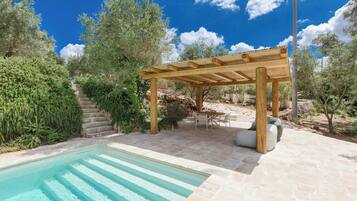 A pool with clear water, a wooden pergola, stone steps, and outdoor furniture.