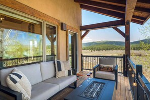 Ein überdachter Außenbereich mit einer Couch und Sesseln, einer Feuerstelle und Blick auf eine bewaldete Landschaft.