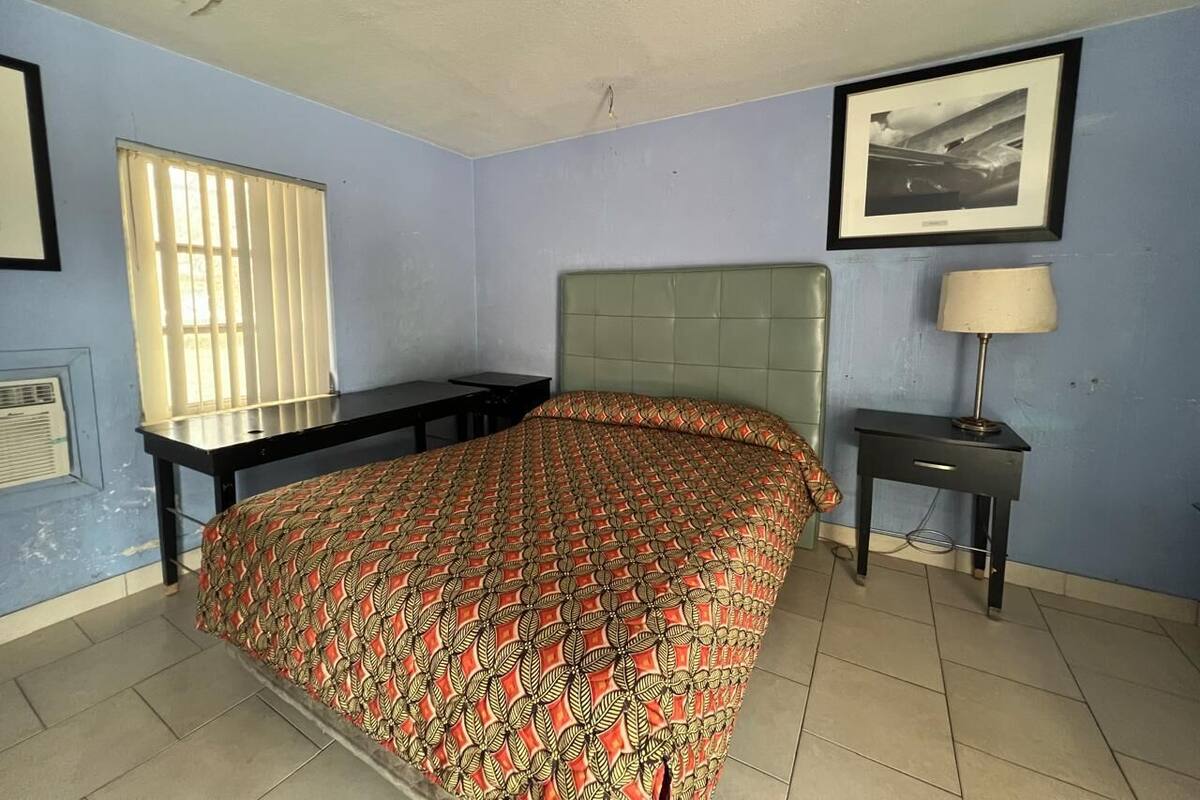 Une chambre à coucher comprenant un lit, une table de chevet, une lampe, une fenêtre avec des stores et un tableau encadré accroché au mur.