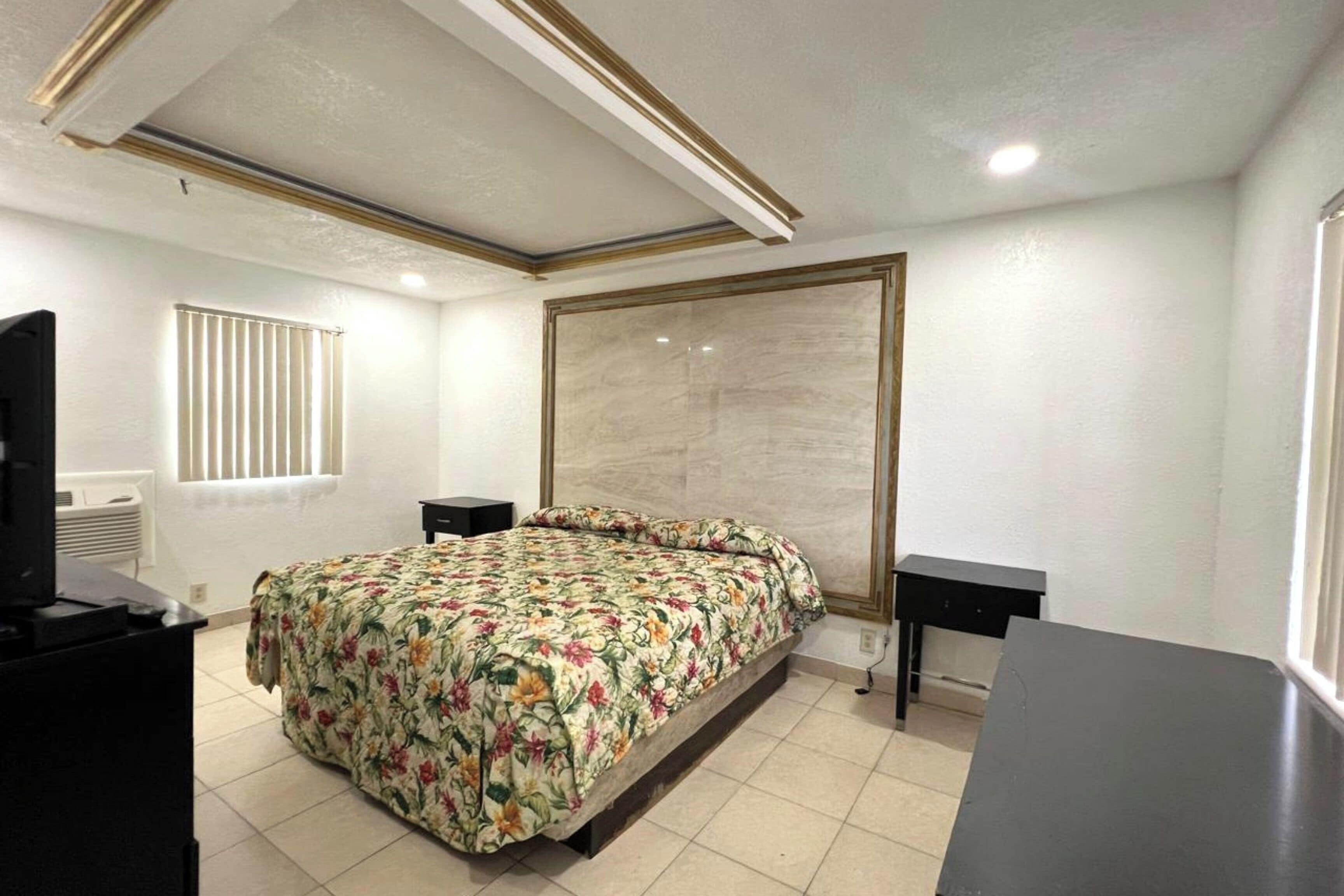 A bedroom with a bed, two bedside tables, a television, and a window with blinds.