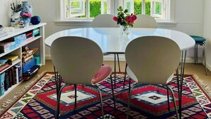 A room with a round table, two chairs, a vase with flowers, a bookshelf, and a view through large windows. - Lovely summerhouse by the beach (Karrebæksminde)