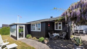 A small, dark-colored building with a glass door entrance, a patio with a table and chairs, and a garden with purple flowers. - Lovely summerhouse by the beach (Karrebæksminde)