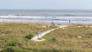 Ein Strand mit einem Pfad zum Wasser, einem Wegweiser und Personen in der Ferne.