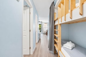 A hallway with a bunk bed, a door, and a view into a room with a bed and a lamp.