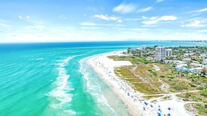 Vista aérea de una playa con una extensa franja de arena blanca, agua turquesa cristalina y un denso grupo de edificios al fondo.