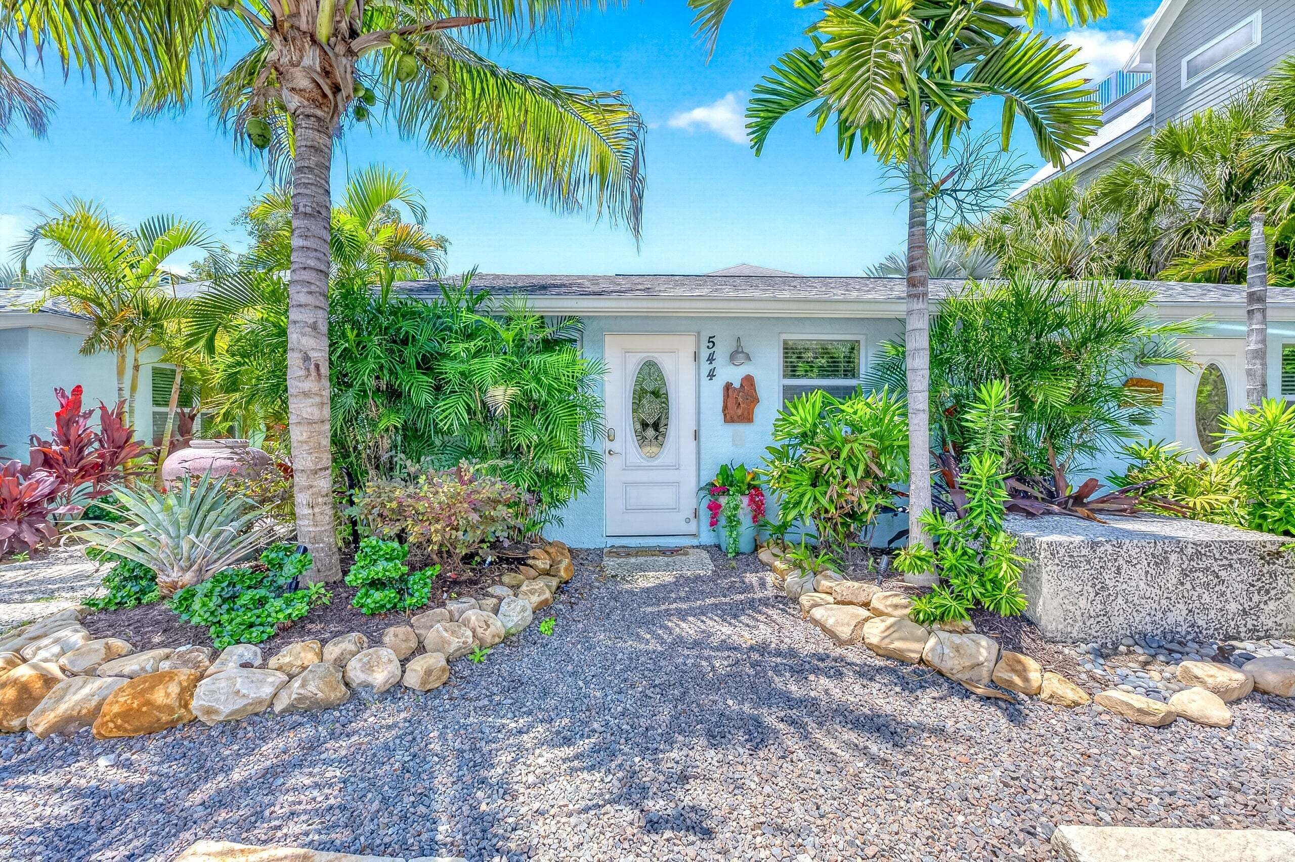 A house with a white door, surrounded by tropical plants and a gravel driveway.