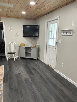 A room with a flat-screen TV mounted on the wall, a white door with a window, and a white chair with a small table.