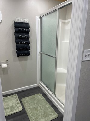 Ein Badezimmer mit Duschabtrennung aus Glas, einem Handtuchhalter mit gefalteten Handtüchern und zwei grünen Teppichen auf dem Boden.