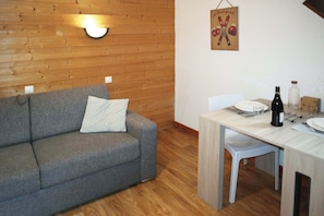 Ein Zimmer mit grauem Sofa, einer Holzwand, einem Esstisch mit Stühlen und einer Wandleuchte.