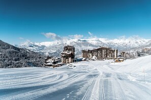 Ein schneebedecktes Skigebiet mit mehreren Gebäuden, darunter ein großes Hotel und kleinere Chalets, vor der Kulisse von schneebedeckten Bergen und blauem Himmel.