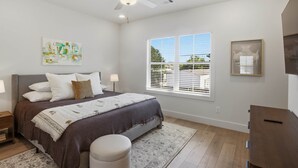 Quarto com cama, mesa de cabeceira, luminária, janela com persianas, ventilador de teto e uma obra de arte na parede.