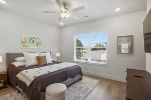 Un dormitorio con cama, mesita de noche, lámpara, ventana con persianas, ventilador de techo y una obra de arte en la pared.