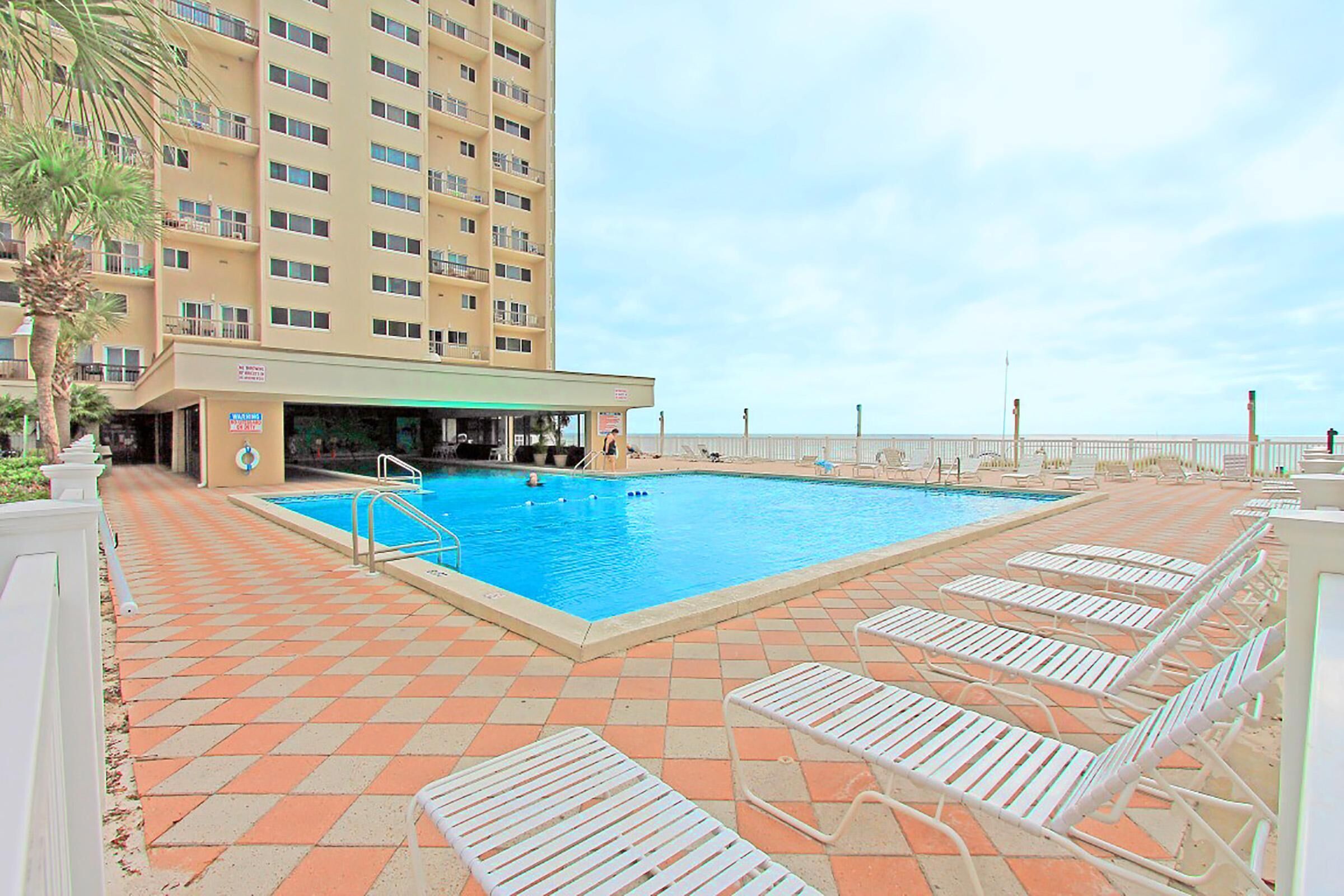 A swimming pool with lounge chairs, a building with multiple floors, and a clear sky.