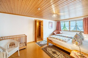 A room with a wooden ceiling, a crib, a bed with patterned bedding, a chair, a window with curtains, and a radiator. - Apartment 'Buche' with Shared Terrace, Shared Garden and Wi-Fi (Sankt Georgen im Schwarzwald)