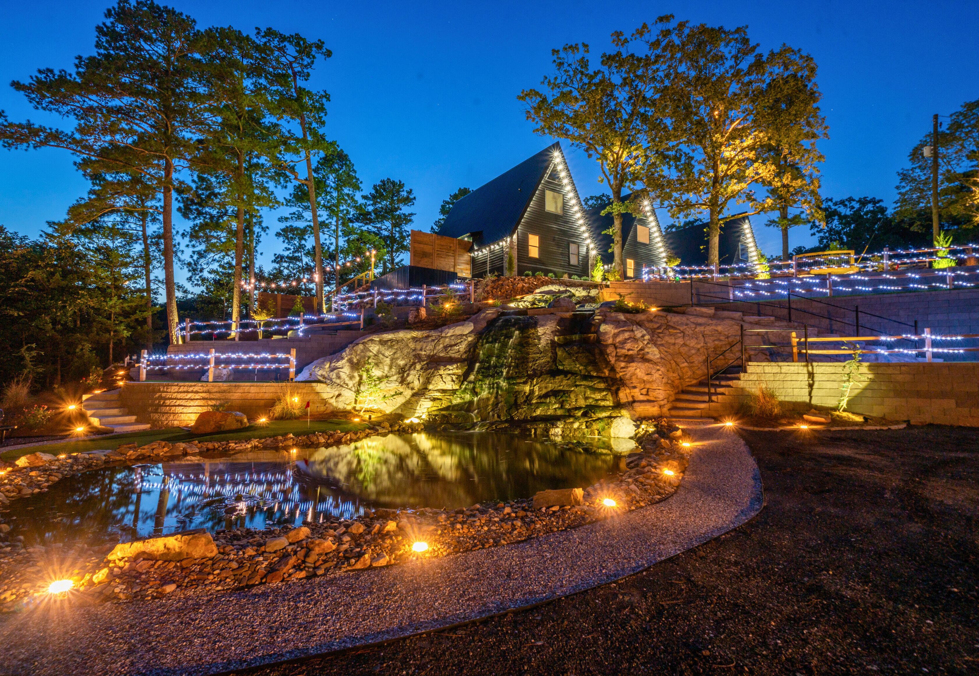 A nighttime view of a property with a pond, illuminated pathways, and a building with a sloped roof.