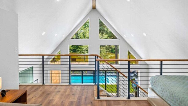 A high-ceilinged room with a wooden floor, a bed, a desk, and a view of a swimming pool through large windows. - Stylish A-Frame Getaway with Pool and Lake Access (Garland County)