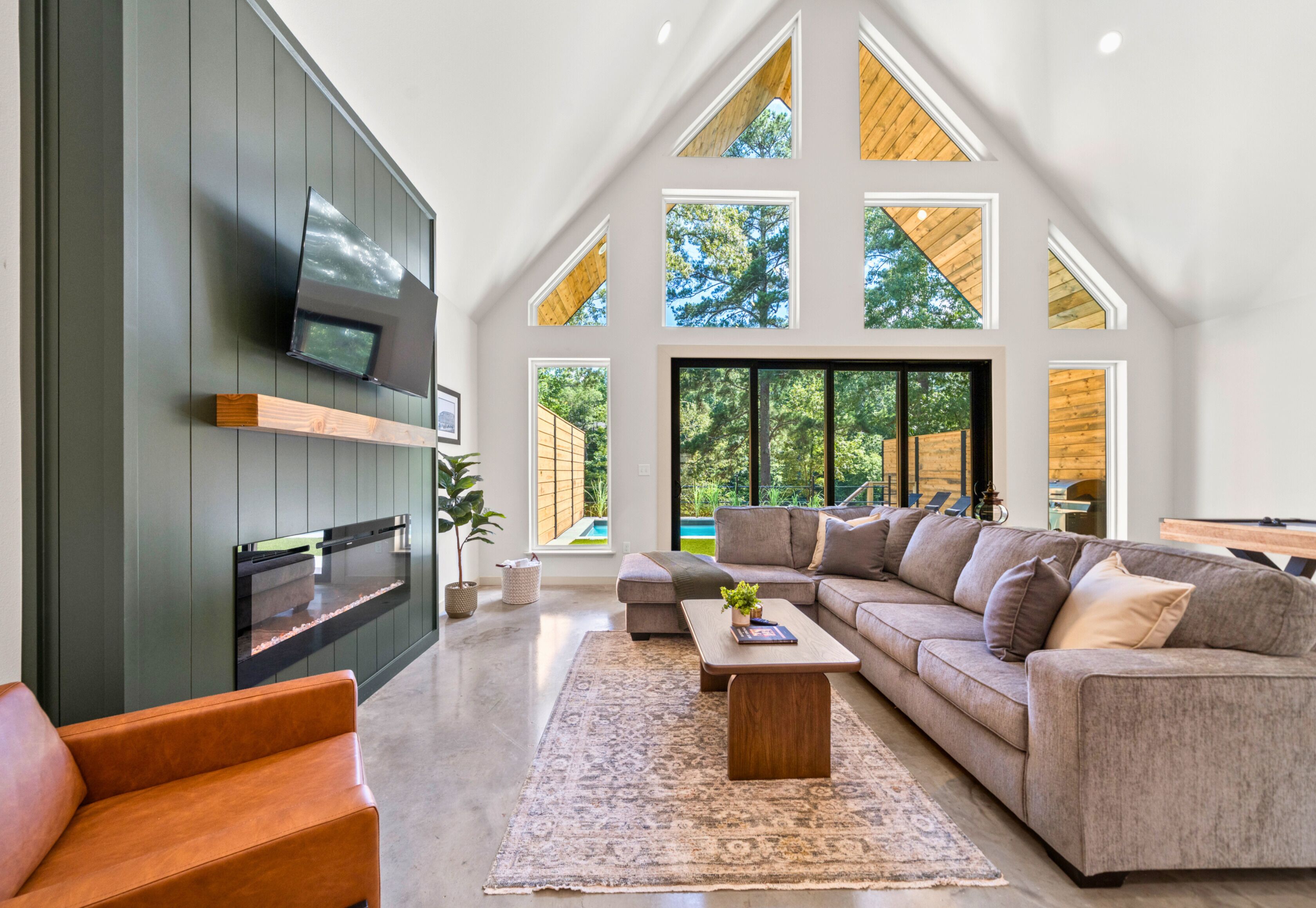 A spacious living room with a large sectional sofa, a flat-screen TV mounted on the wall, and a large window offering a view of the outdoors.