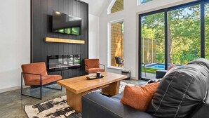 A modern living room with a flat-screen TV, a wooden coffee table, leather sofas, and a large window with a view of greenery. - 3 Side-By-Side Modern Lakefront A-Frame Cabins (Garland County)
