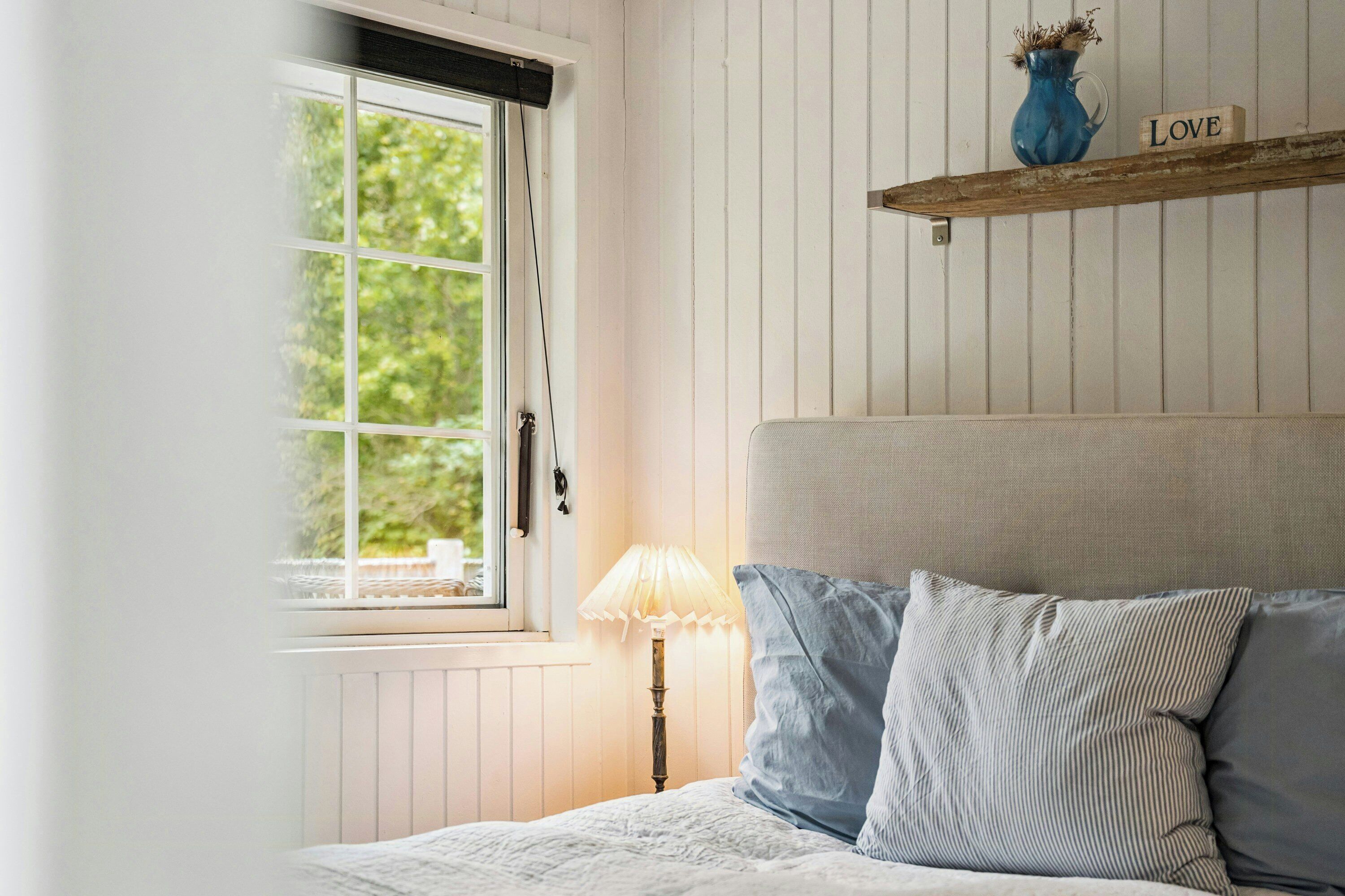 Une chambre à coucher avec un lit, une fenêtre, une lampe et une étagère sur laquelle se trouvent un vase et un bloc portant le mot « AMOUR ».
