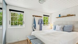 Een slaapkamer met een groot bed, een houten bankje, twee ramen met gordijnen en een plank met decoratieve objecten.