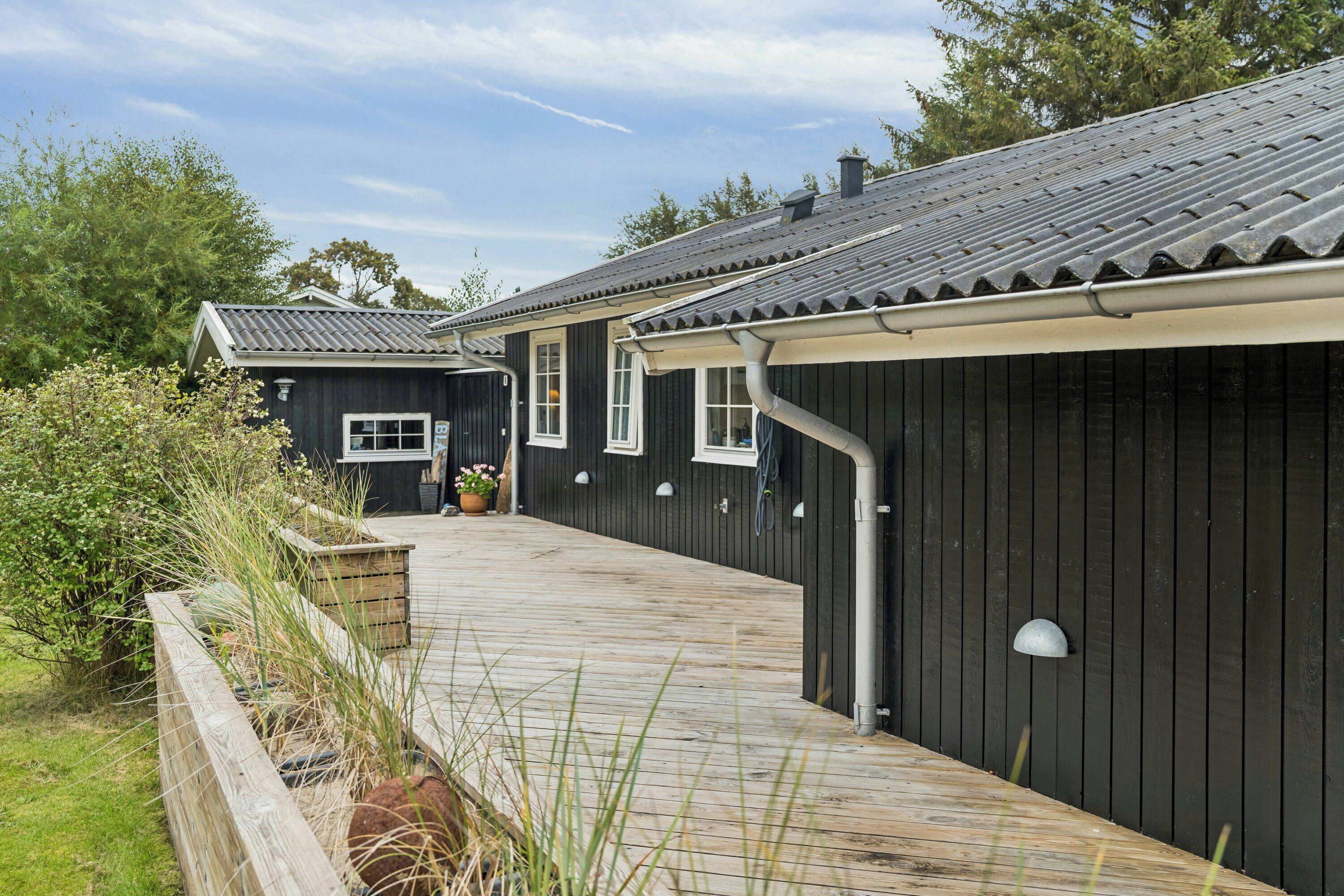 A wooden deck leading to a dark wooden building with a tiled roof, surrounded by greenery.