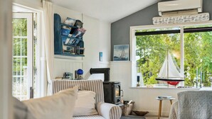 Een gezellige woonkamer met een bank, een klein tafeltje, een boekenkast, een open haard en een groot raam met uitzicht op groen.