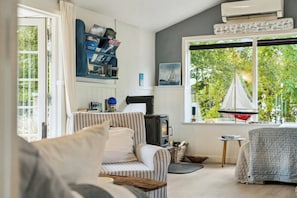 Ein gemütliches Wohnzimmer mit Sofa, einem kleinen Tisch, einem Bücherregal, einem Kamin und einem großen Fenster mit Blick ins Grüne.