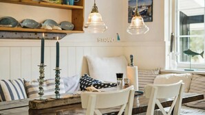 Een gezellig eetgedeelte met een houten tafel, witte stoelen en een bank. Er hangen twee pendelverlichting, een houten plank met decoratieve borden, en een raam met uitzicht op buiten.