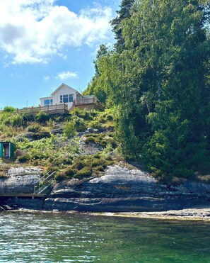 Ein Küstenhaus mit Holzterrasse, direkt an einer felsigen Küstenlinie gelegen, mit klarem Wasser im Vordergrund.