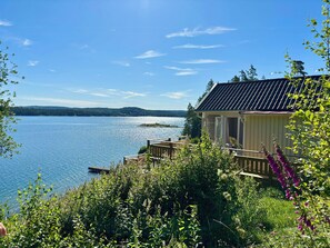 Eine Blockhütte am Seeufer mit einer Terrasse, umgeben von üppiger Vegetation und freiem Blick auf den See.