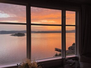 Ein Zimmer mit großem Fenster mit Blick auf ruhiges Wasser bei Sonnenuntergang.
