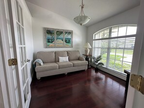 Una habitación con piso de madera, un sofá color beige, un cuadro, una lámpara y una ventana con persianas.