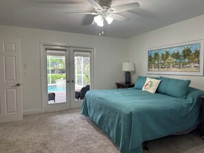 A bedroom with a bed, a ceiling fan, a painting, and a view of a pool through a door.