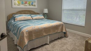 Un dormitorio con cama, ventilador de techo, ventana con persianas y un cuadro con temática playera en la pared.