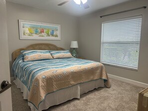 Un dormitorio con cama, ventilador de techo, ventana con persianas y un cuadro con temática playera en la pared.