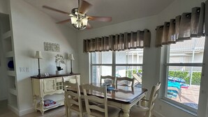 Un comedor con mesa y sillas, ventilador de techo, ventana con cortinas y vista al exterior.