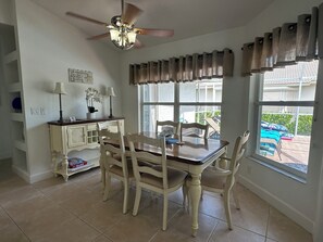 Un comedor con mesa y sillas, ventilador de techo, ventana con cortinas y vista al exterior.