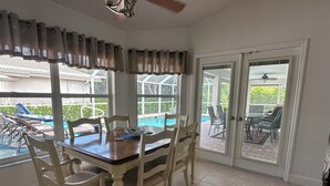 Comedor con mesa y sillas, ventilador de techo y vista a una piscina a través de amplios ventanales.