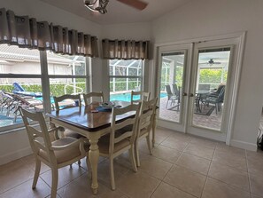 Comedor con mesa y sillas, ventilador de techo y vista a una piscina a través de amplios ventanales.