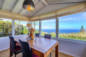 A dining area with a wooden table and chairs, a vase with dried flowers, and a view of the ocean.