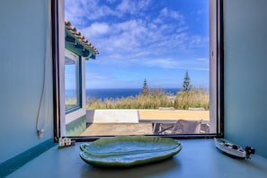 A window with a view of the ocean and a patio area with a green dish and a small boat model.