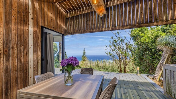A wooden deck with a table and chairs, a glass door leading outside, and a view of the ocean. - GuestReady - Casa do Horizonte (Mosteiros)