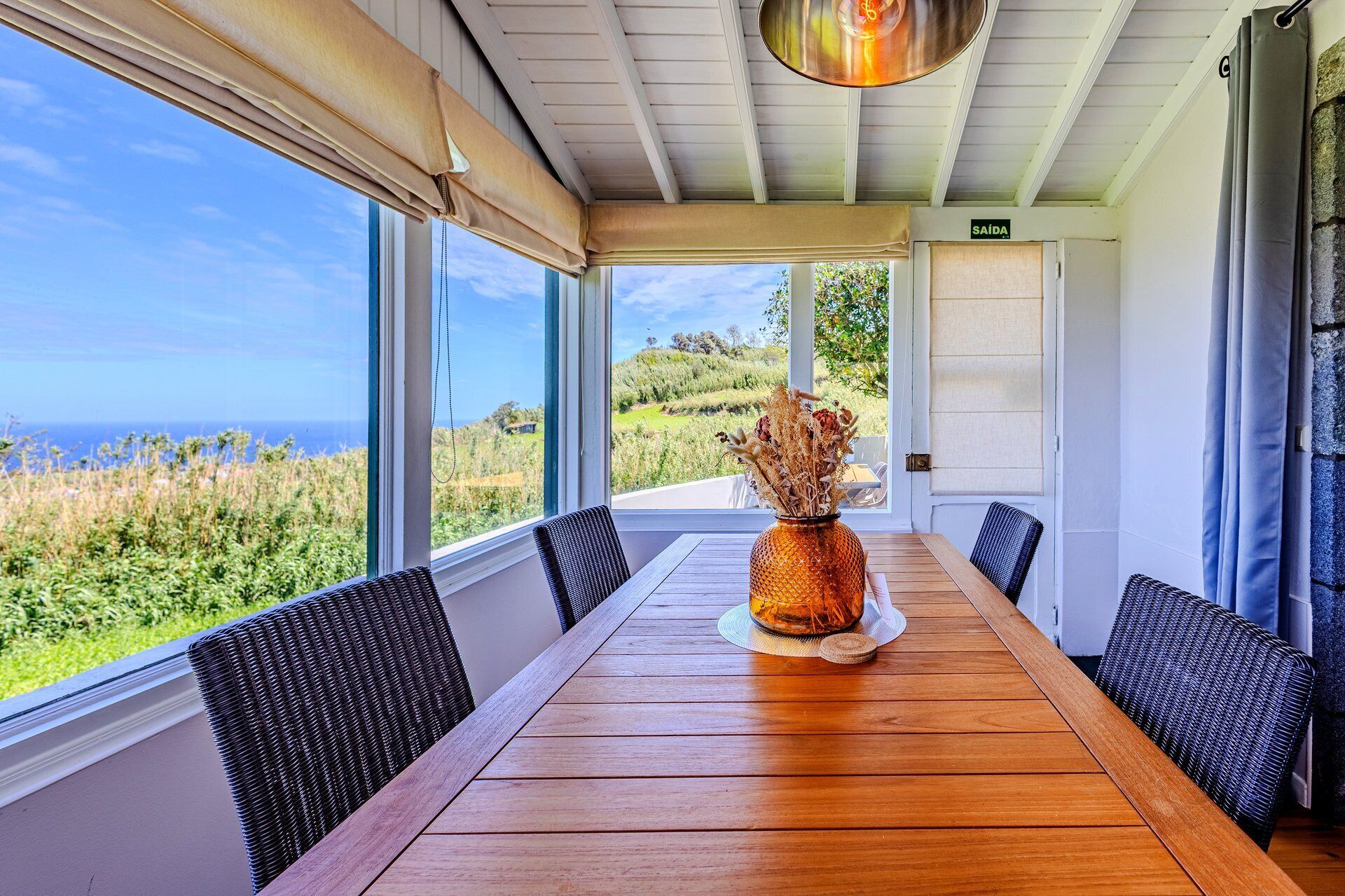 A dining area with a wooden table, chairs, a vase with dried flowers, and a view of the ocean.