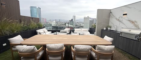 A rooftop patio with a wooden table and chairs, a grill, and a city view.