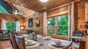 Interior de una cabaña de madera con una mesa de comedor para cuatro, un sofá y una ventana con vistas a la vegetación.